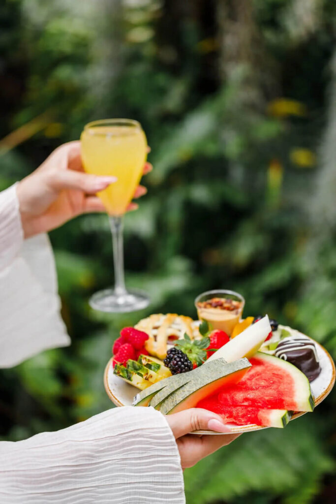 A person holding a platter of fruit including watermelon and berries, with a glass of drink in the other hand